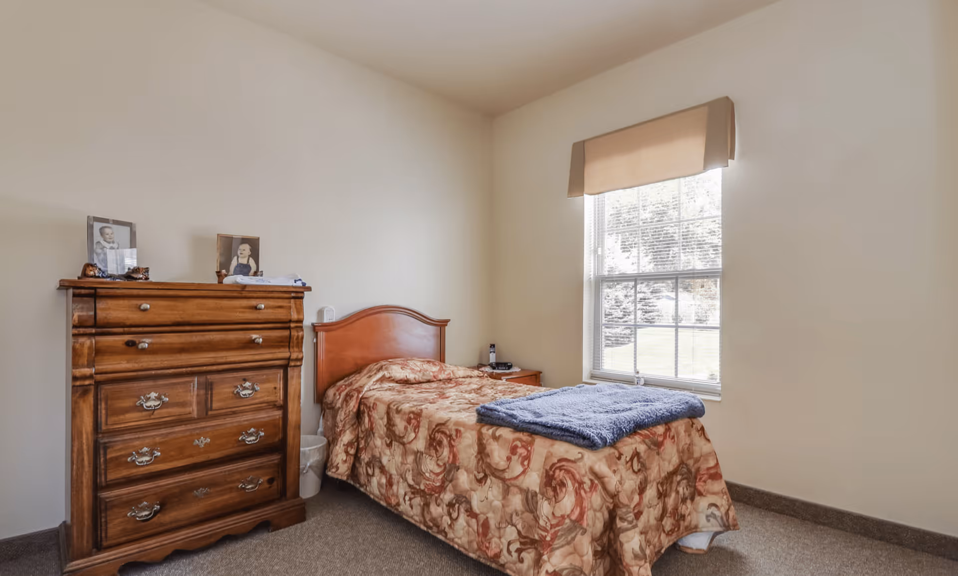 A small bedroom with a single bed covered with a patterned bedspread and a folded blue blanket at the foot. Next to the bed is a wooden nightstand with a phone and other small items. A large window with a beige valance lets in natural light. On the left side of the room is a wooden dresser with framed photos on top. The walls are plain and light-colored, and the floor is carpeted.