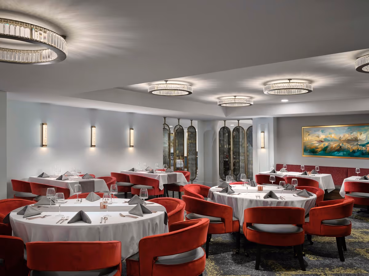 Elegant dining room with round and rectangular tables covered in white tablecloths, set with gray napkins, glassware, and cutlery. The room features red upholstered chairs, modern circular ceiling lights, wall sconces, a decorative wine display, and a colorful abstract painting on the wall.