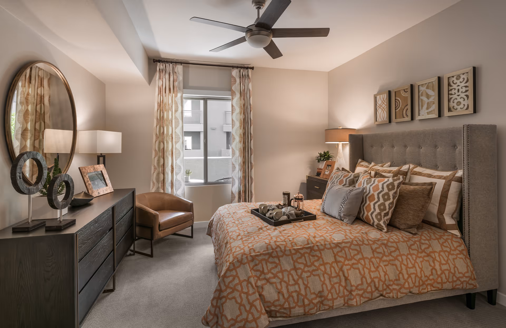A cozy bedroom with a large bed covered in patterned orange and beige bedding and multiple pillows. There is a dark wooden dresser with decorative items and a round mirror on the left side. A brown leather chair is positioned near a window with patterned curtains. On the right side, there is a nightstand with a lamp and framed pictures. The walls are decorated with four framed abstract art pieces above the bed. The room has a ceiling fan and neutral-colored walls and carpet.