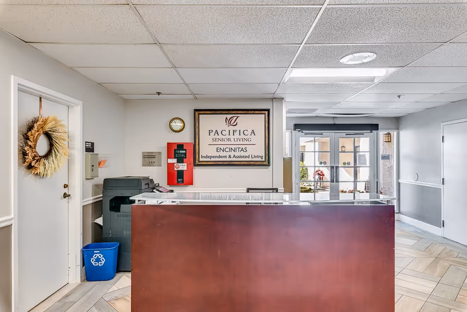 Reception area of Pacifica Senior Living Encinitas with a wooden front desk, a framed sign on the wall, a clock, a fire alarm, a printer, a recycling bin, and a door decorated with a wreath. Glass doors lead to an outdoor area in the background.