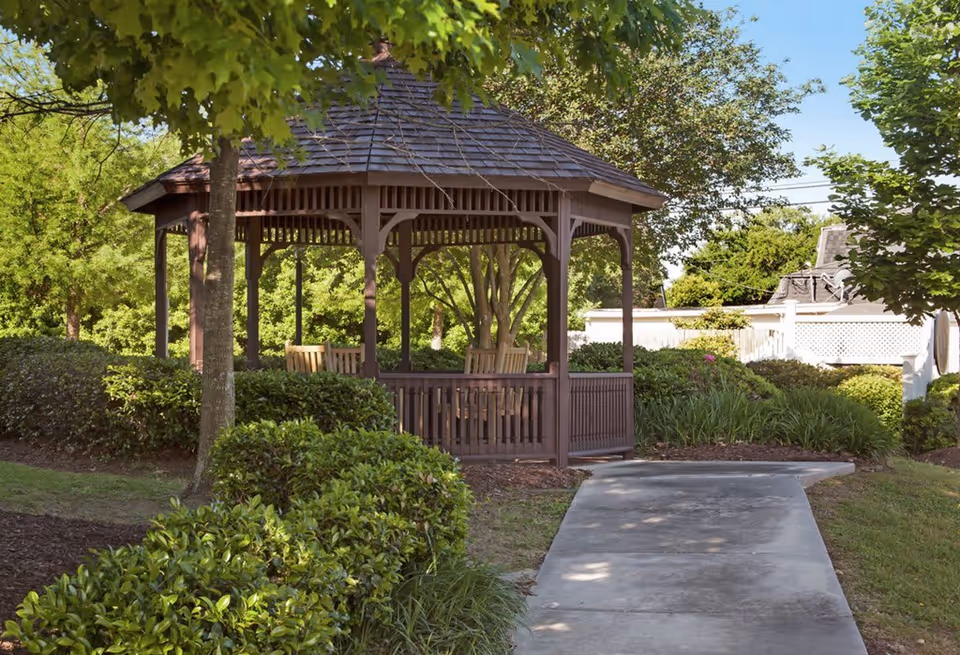 A wooden gazebo with a shingled roof surrounded by green bushes and trees. There is a concrete pathway leading up to the gazebo, which contains wooden chairs inside. The scene is outdoors with clear skies and lush greenery.