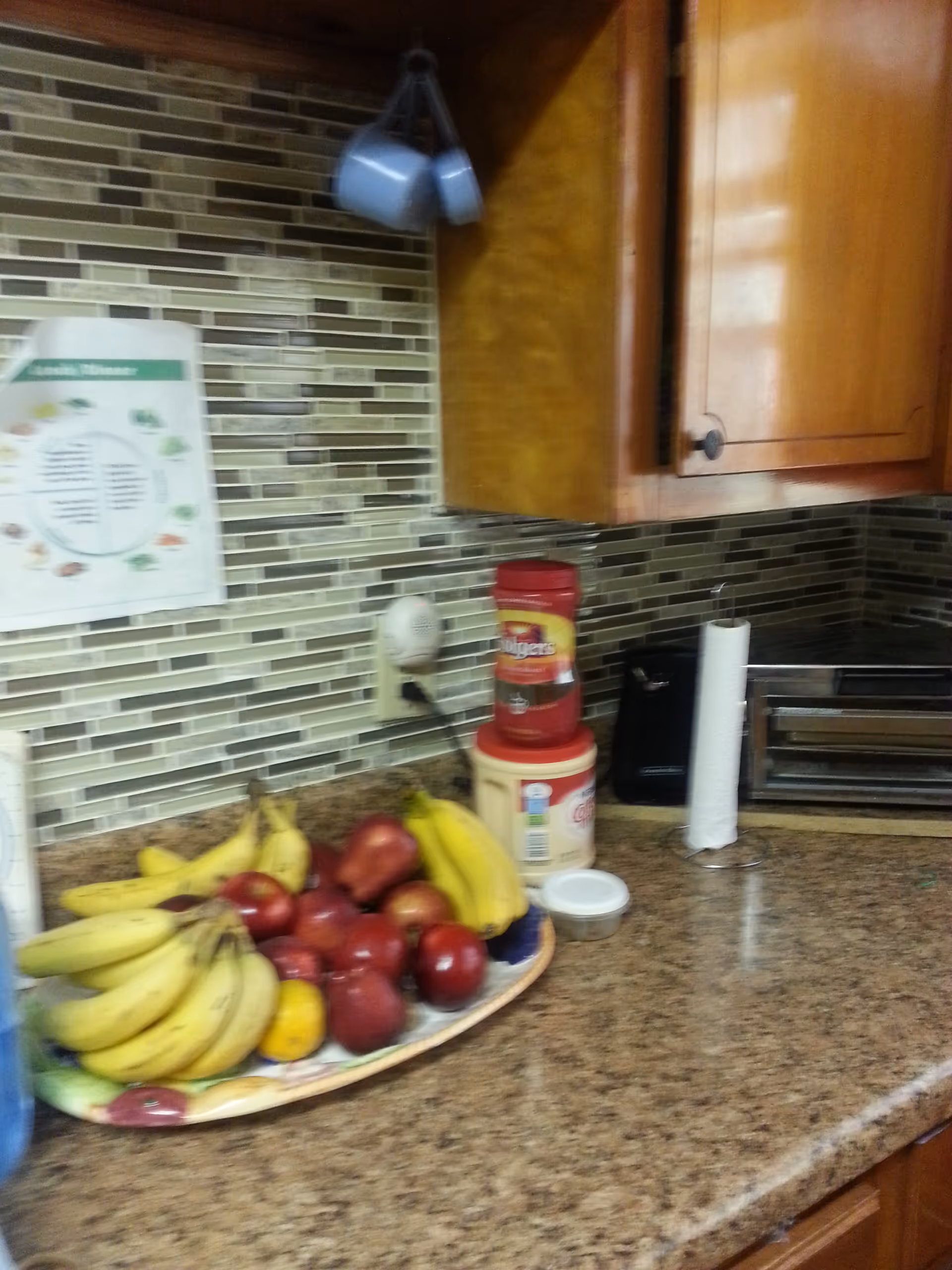 Kitchen countertop with a bowl of fruit (bananas and apples), paper towel holder, cabinets and a mosaic tile backsplash.