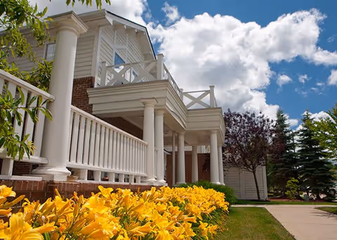 Exterior view of a building with white columns and railings, a balcony, and a brick base. Yellow flowers and green grass are in the foreground, with trees and a partly cloudy blue sky in the background.