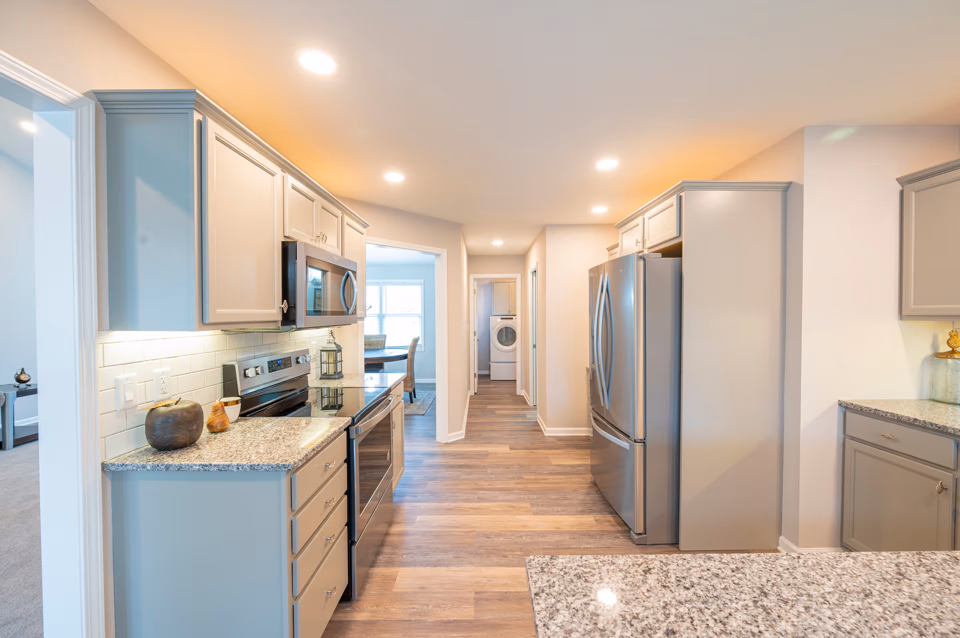 Bright modern kitchen with gray cabinets, granite countertops, stainless steel appliances including a refrigerator, stove, and microwave. The kitchen has wood flooring and recessed ceiling lights. A hallway leads to a laundry area with a washer and dryer visible in the background.