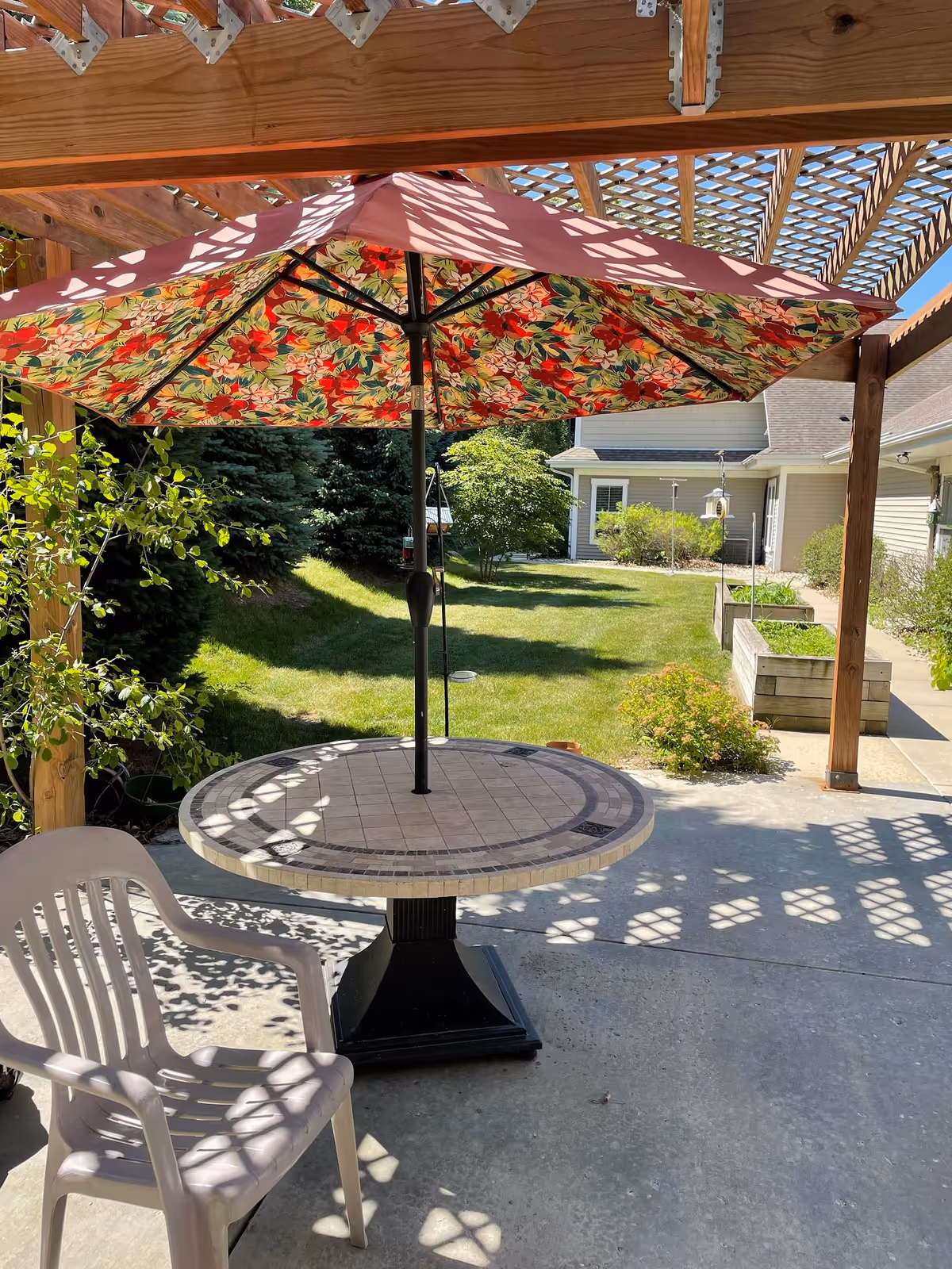 Outdoor patio area with a round tiled table and a floral-patterned umbrella providing shade. A white plastic chair is positioned next to the table. The patio is covered by a wooden pergola casting patterned shadows on the concrete floor. In the background, there is a grassy lawn, some bushes, raised garden beds, and a light-colored building.