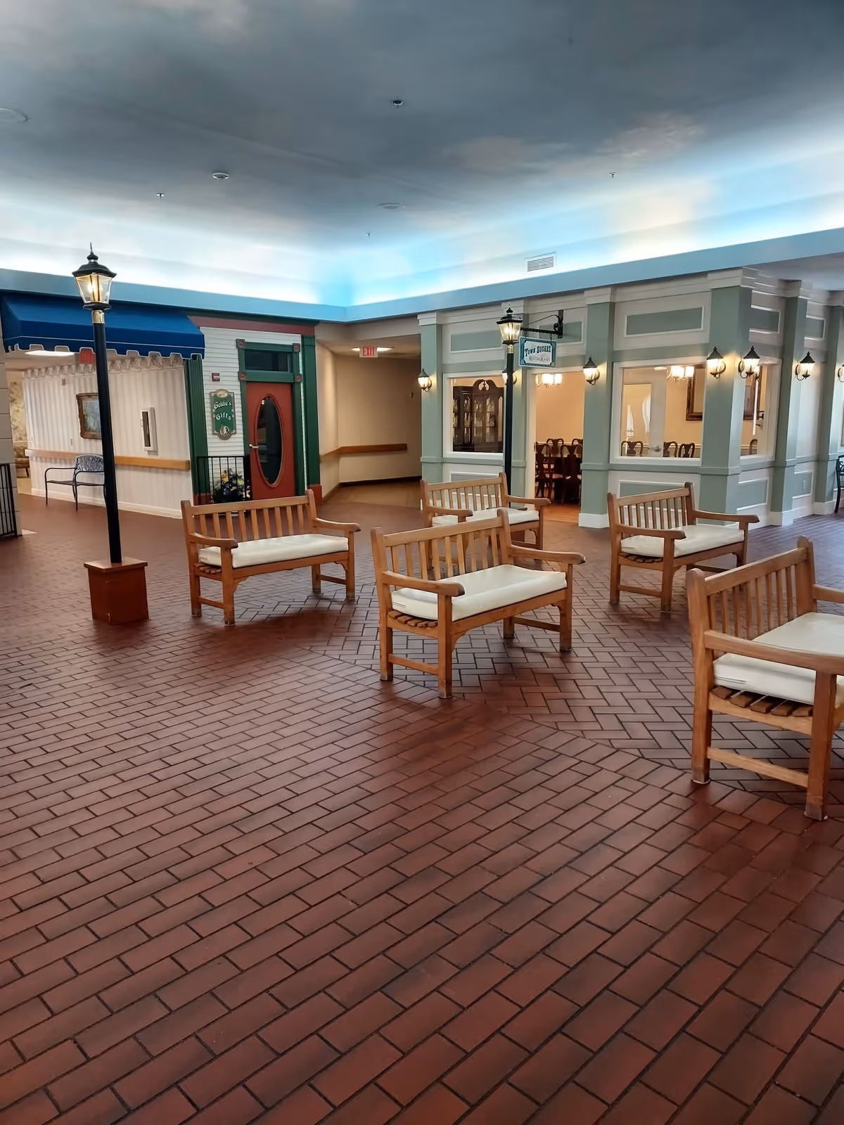 Indoor seating area with wooden benches that have white cushions, surrounded by streetlamp-style lights. The floor is covered with reddish-brown tiles. The background shows a hallway with a red door and a green-framed window, and a room with dining tables and chairs visible through large windows.