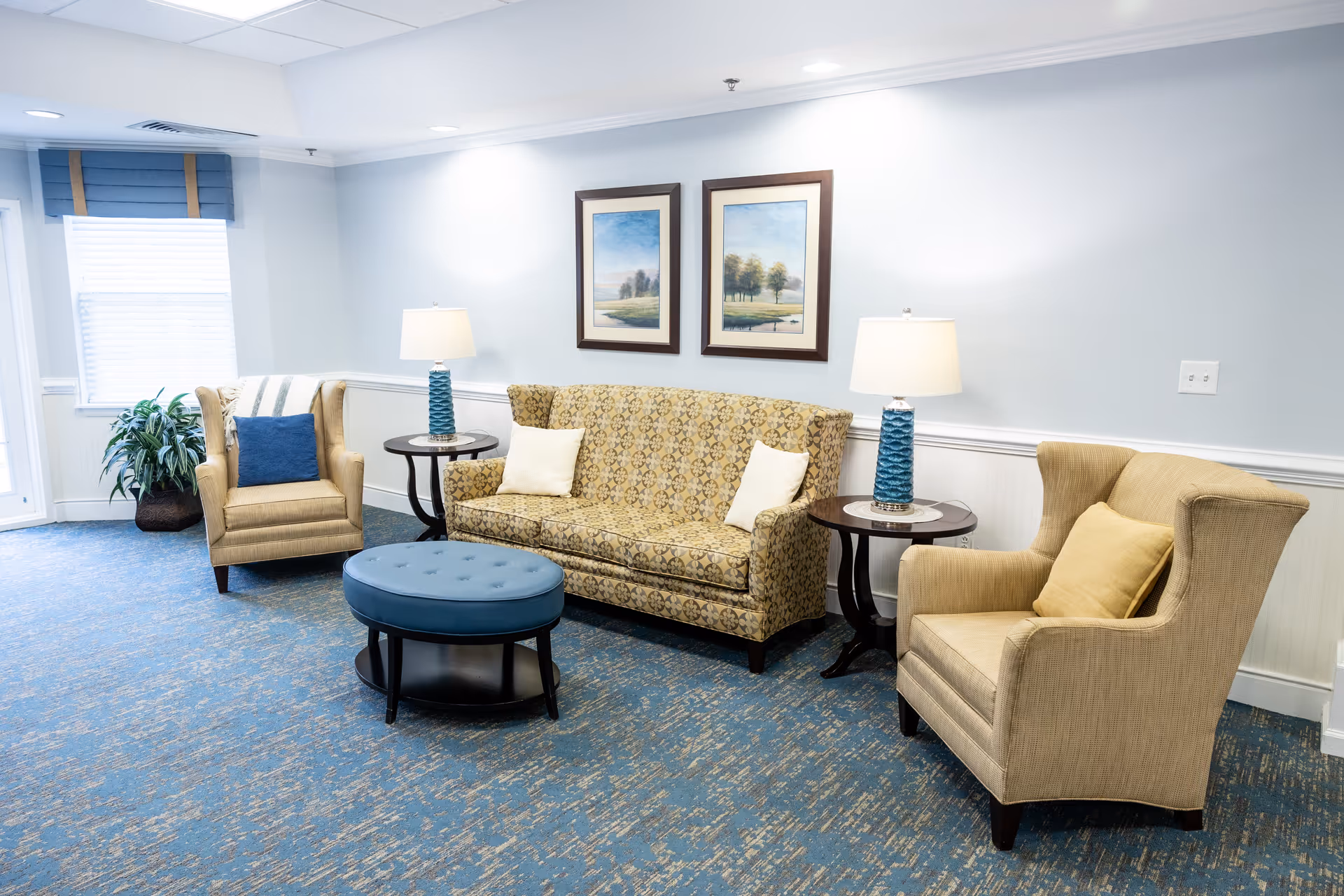A bright common area with a patterned sofa, two upholstered armchairs, a round blue ottoman, side tables with lamps, framed artwork, and a window.