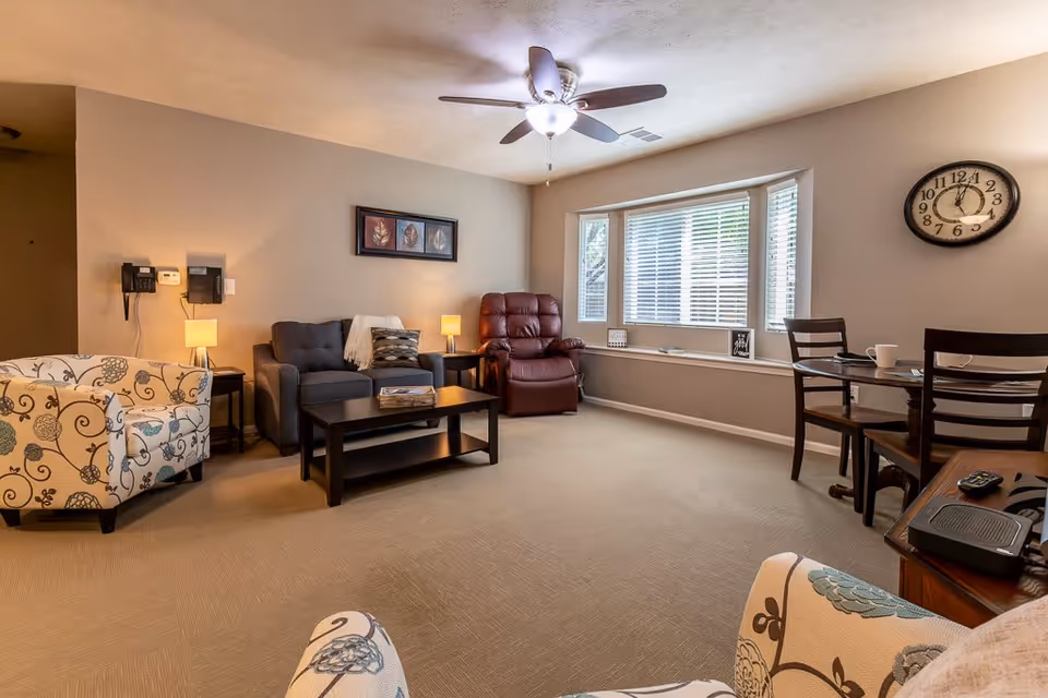 A cozy living room in an assisted living facility featuring a patterned armchair in the foreground, a dark gray loveseat with a throw blanket and pillow, a maroon recliner, a wooden coffee table, and a small dining table with two chairs near a large window with blinds. The room is softly lit by two table lamps and a ceiling fan with a light. A wall clock and framed artwork are also visible.