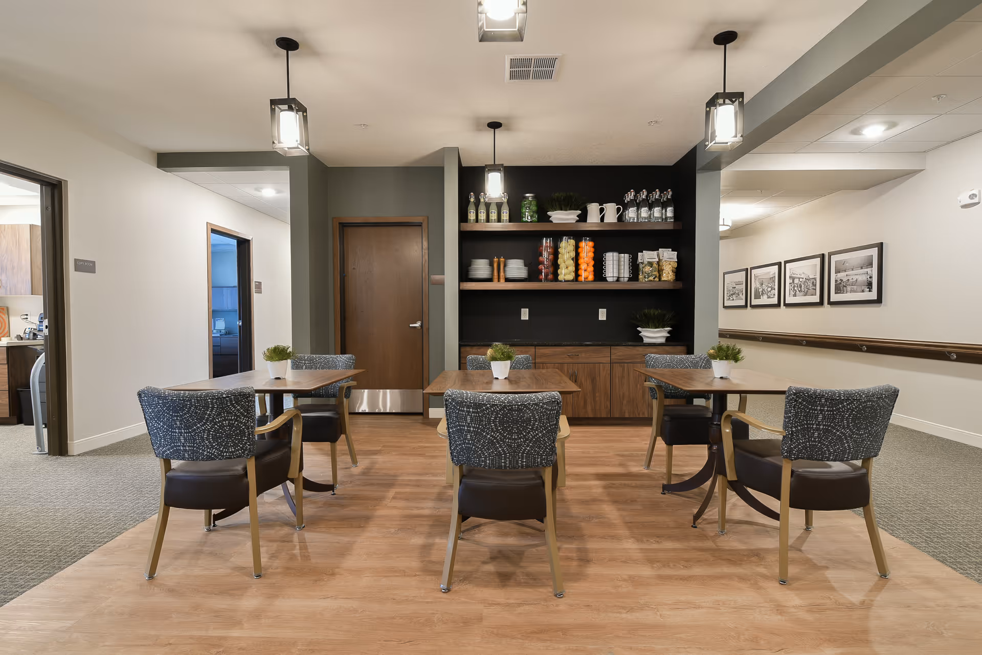 Communal dining area with four tables and upholstered chairs, a shelving unit stocked with dishes and jars, and a central doorway.