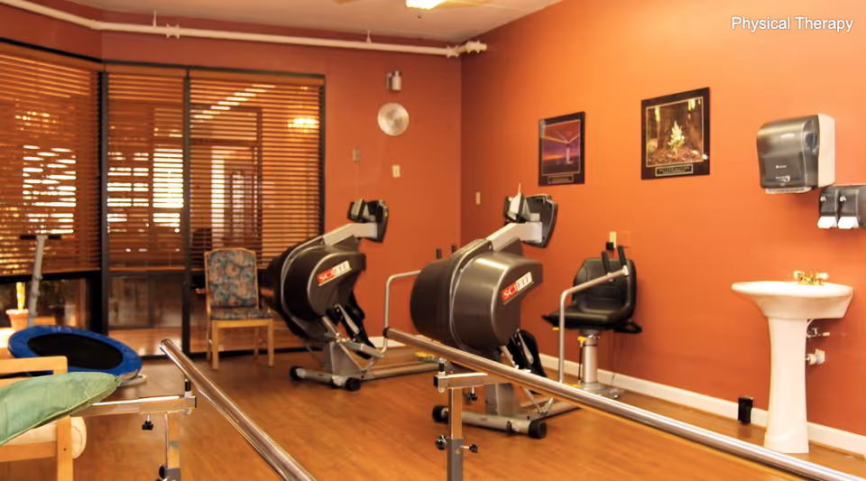 Physical therapy room with two exercise machines, parallel bars for walking support, a chair, a sink with soap dispenser, and two framed pictures on the orange wall.