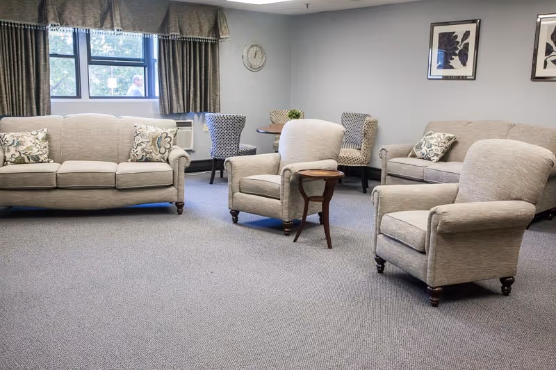A senior living center common area with two beige sofas and two beige armchairs arranged on a carpeted floor. There is a small wooden side table between the armchairs. In the background, there is a round table with four patterned chairs, a window with curtains, a wall clock, and two framed pictures on the wall.