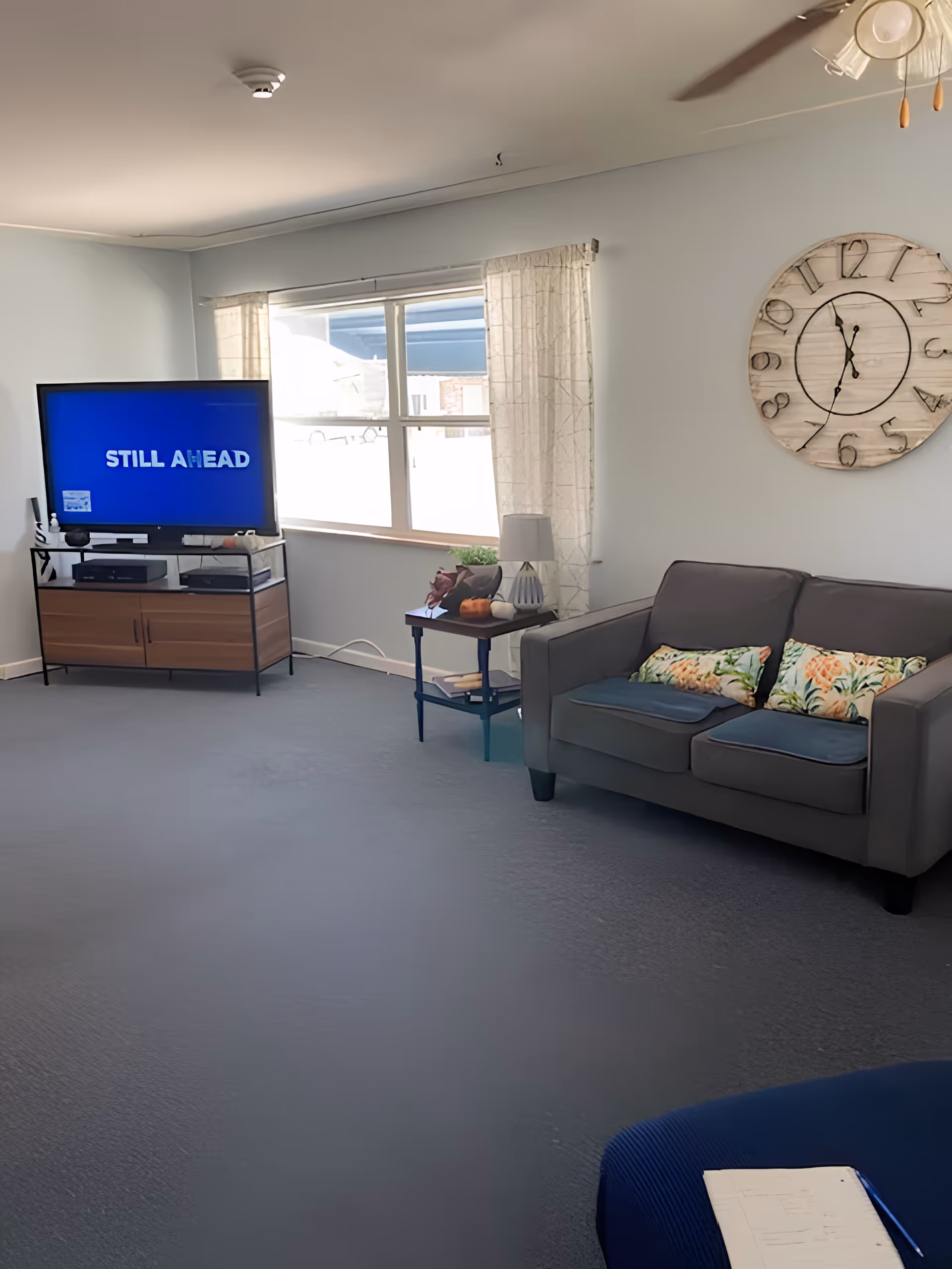 A bright living room with a TV on a wooden stand, a window with curtains, a small side table, a gray loveseat with patterned pillows, and a large wall clock.