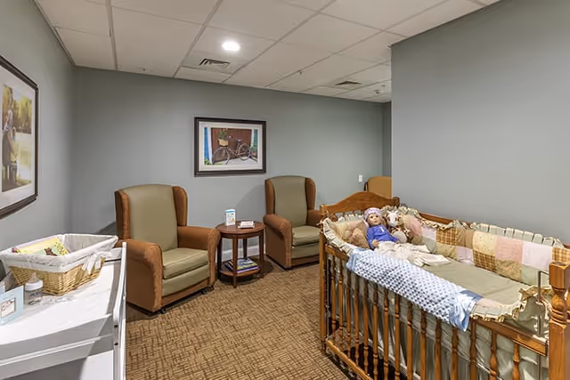 A cozy room with two brown armchairs and a small round wooden table between them. There is a wooden crib with a doll lying inside, covered with a patchwork quilt. On the left side, there is a white changing table with a basket and baby supplies. The walls are painted light gray and decorated with framed pictures. The floor is carpeted with a patterned design.