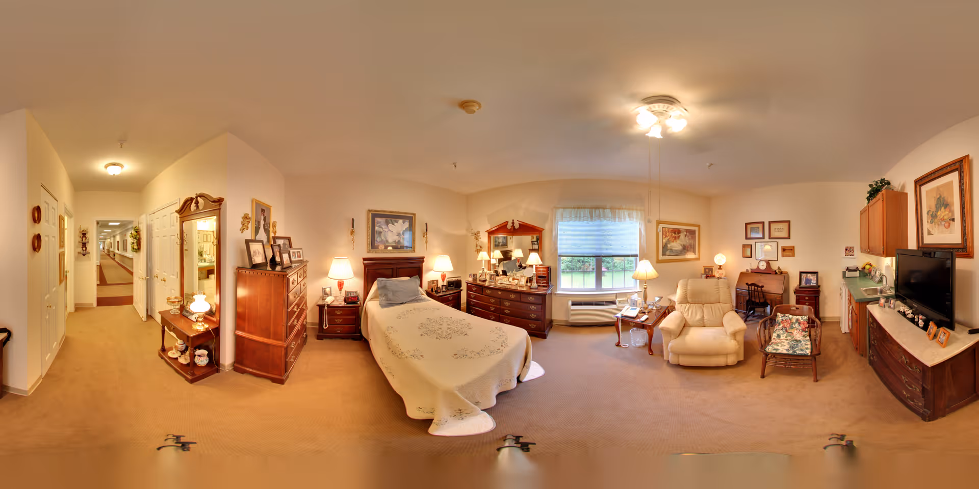 A cozy and well-lit bedroom in a senior living facility featuring a single bed with a white quilt, wooden dressers, a comfortable armchair, a small table with a lamp, a television on a wooden cabinet, framed artwork on the walls, and a window with a view of greenery outside.