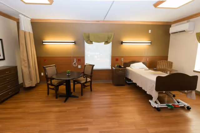 A senior living facility room with a single hospital-style bed on the right side, a small bedside table, a round table with three chairs in the center, wooden flooring, and a window with a green valance in the middle of the back wall. The room has warm lighting and an air conditioning unit mounted on the wall.