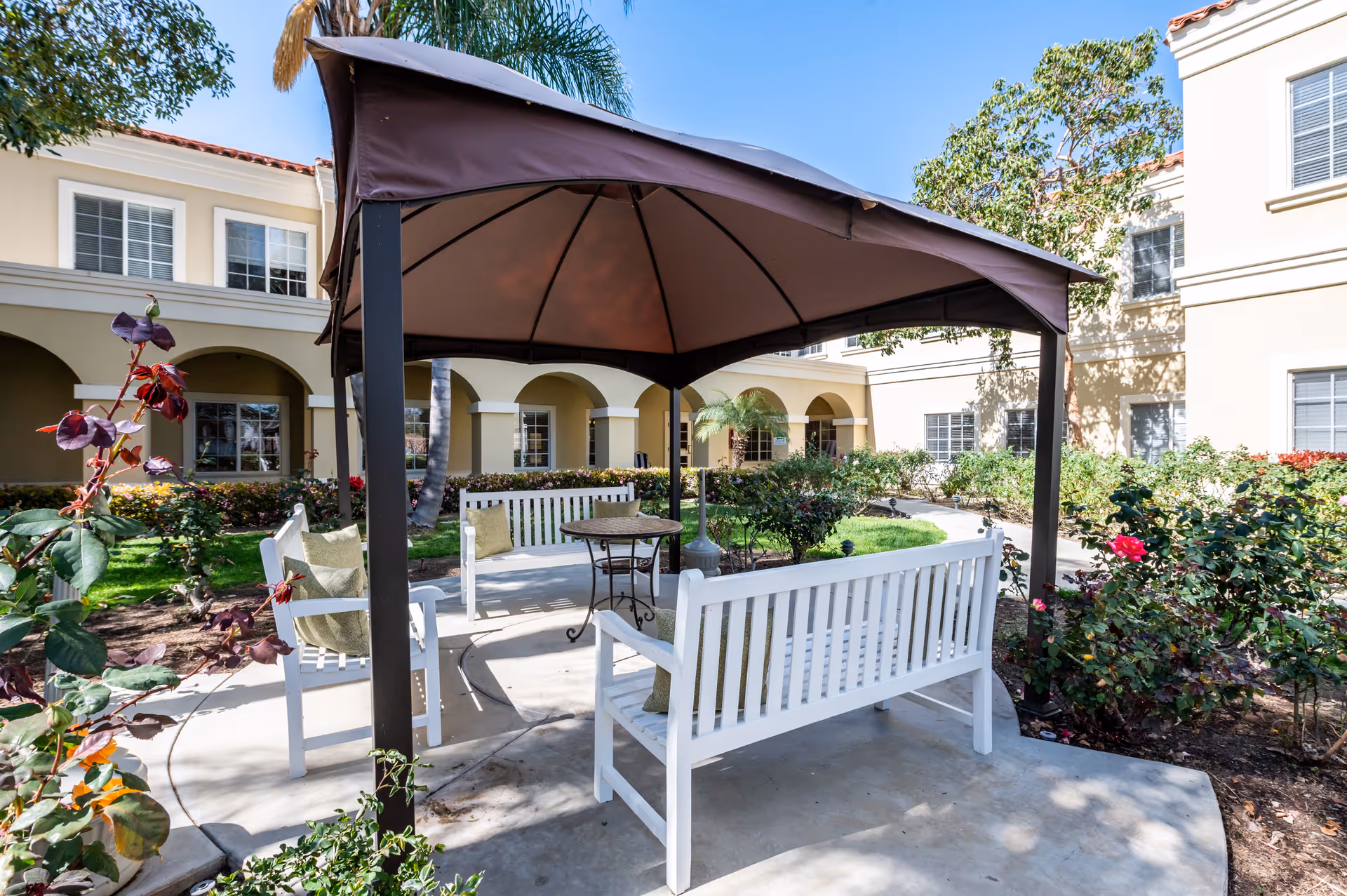 Outdoor seating area with white benches and a small round table under a brown canopy, surrounded by garden plants and flowers, with a beige building featuring arched windows in the background.