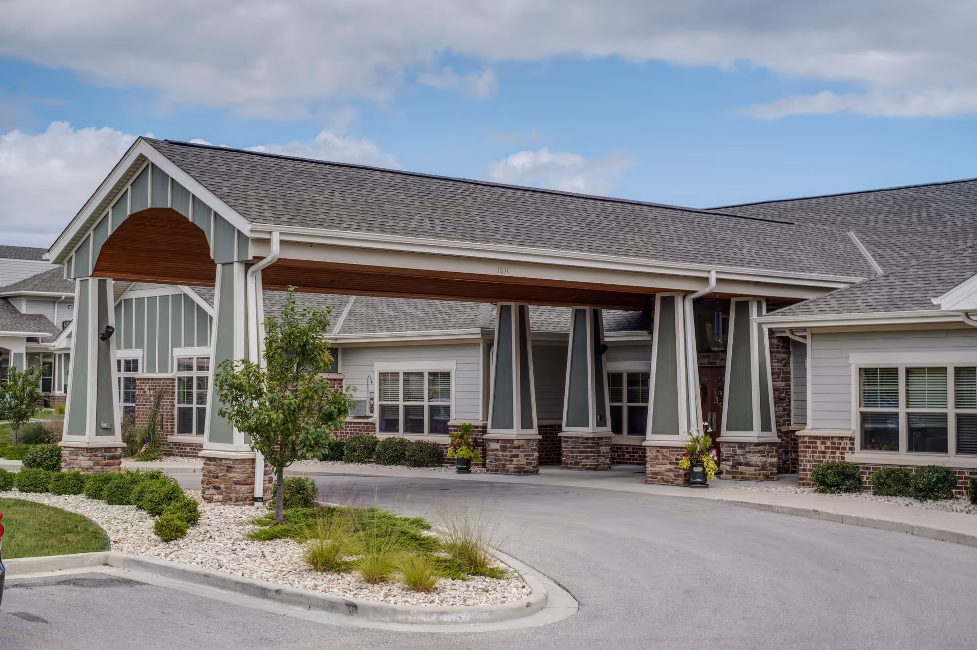 Exterior view of Heritage Court Memory Care facility showing the covered entrance with pillars, landscaped greenery, and a curved driveway under a partly cloudy sky.