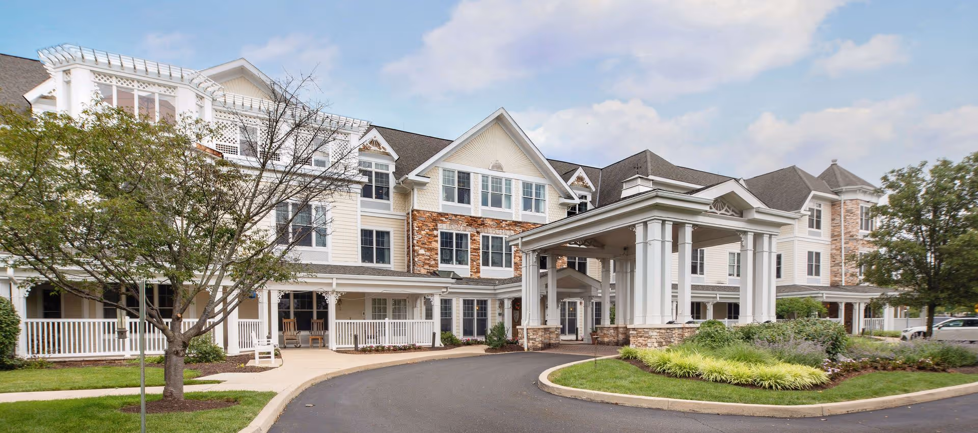 Front exterior of a three-story senior living facility with a covered porte-cochere, wraparound porch, and landscaped driveway.