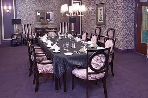 A formal dining room with a long rectangular table covered with a dark tablecloth. The table is set with white napkins, plates, glasses, and silverware. The room features purple carpet, patterned wallpaper, and several upholstered chairs with a pink and white geometric pattern. There are two table lamps on side tables and a chandelier hanging above the table. A framed picture and a mirror are on the walls.