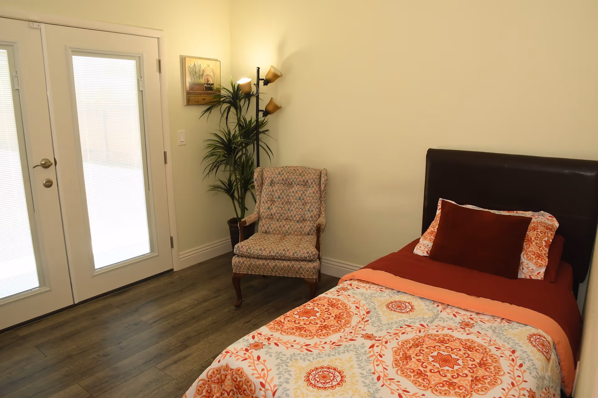 A bedroom with a single bed featuring a patterned orange and white bedspread and a dark brown headboard. Next to the bed is a cushioned armchair with a patterned fabric. There is a tall floor lamp with three lights and a potted plant beside it. The room has wooden flooring and a set of glass double doors with blinds, letting in natural light. A framed picture hangs on the wall above the chair.