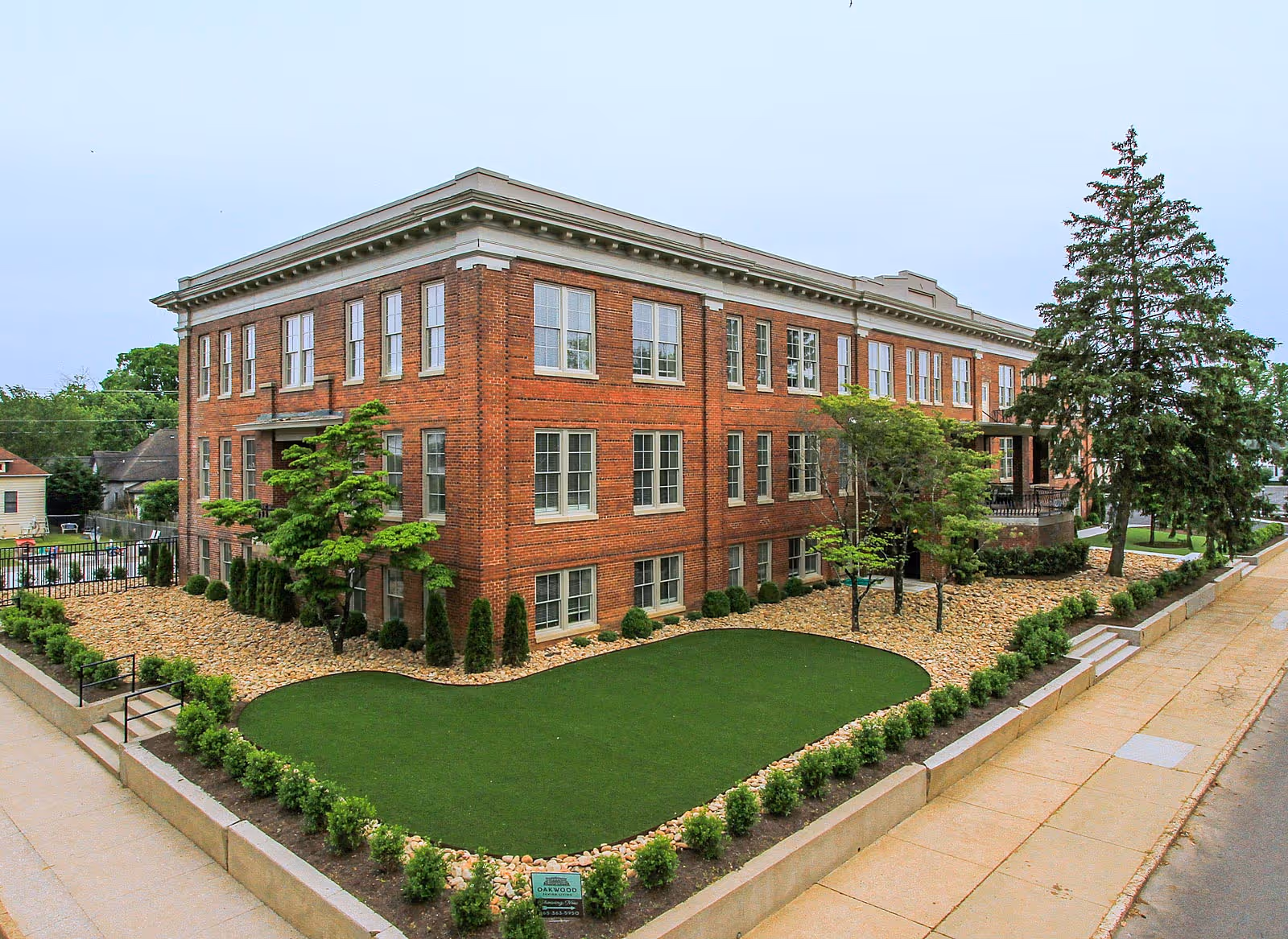 Exterior view of a large, three-story brick building with many windows, surrounded by landscaped grounds including a green lawn, small trees, and shrubs. A sidewalk runs alongside the building, and there is a sign in front that reads Oakwood Senior Living.