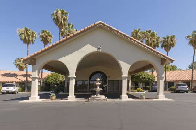 Front exterior view of a single-story building with a large covered entrance supported by columns, a three-tiered fountain in the center, palm trees in the background, and parked cars on either side under a clear blue sky.