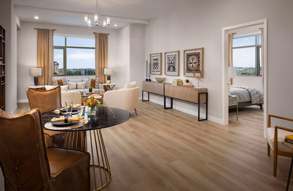 A bright and modern living and dining area with large windows showing an outdoor view. The dining area features a round black marble table set with plates, glasses, and napkins, surrounded by brown leather chairs. The living area has beige armchairs and a sofa with cushions, flanked by two table lamps on side tables. On the right wall, there is a long wooden console table with decorative items and three framed artworks above it. A doorway leads to a bedroom with a bed, nightstand, and window with a similar outdoor view. The flooring is light wood, and the walls are painted white.