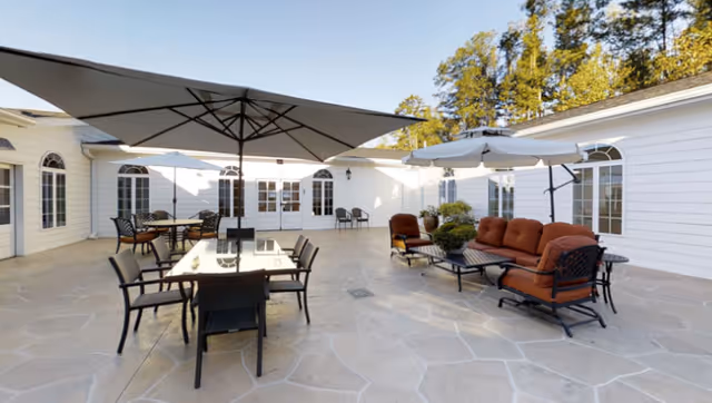 Outdoor patio area with stone flooring, featuring multiple seating arrangements including tables with umbrellas and cushioned chairs. The space is surrounded by white buildings with large windows and doors, and trees are visible in the background under a clear sky.