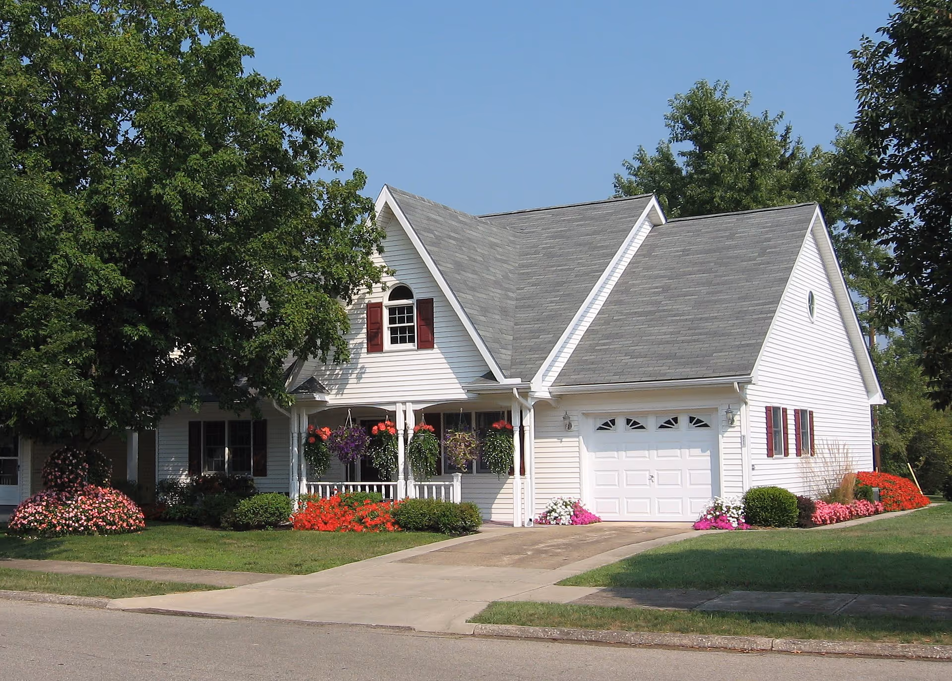 A white house with a gray shingled roof, a garage, and a front porch decorated with hanging flower baskets. The house is surrounded by green grass, colorful flower beds, and large trees under a clear blue sky.