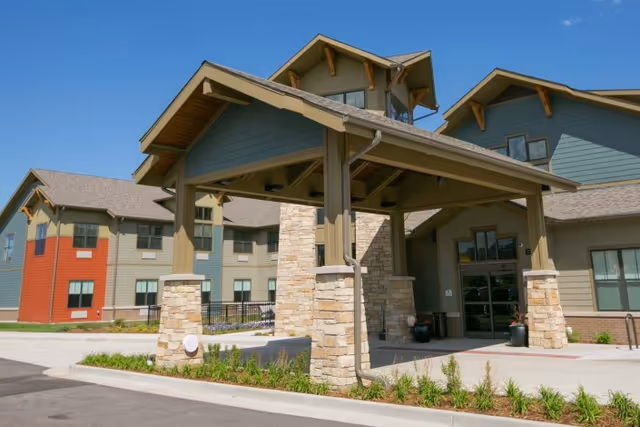 Exterior view of The Healthcare Resort Of Colorado Springs showing the main entrance with a covered drop-off area supported by stone pillars, surrounded by a multi-story building with a mix of beige, green, and red siding under a clear blue sky.