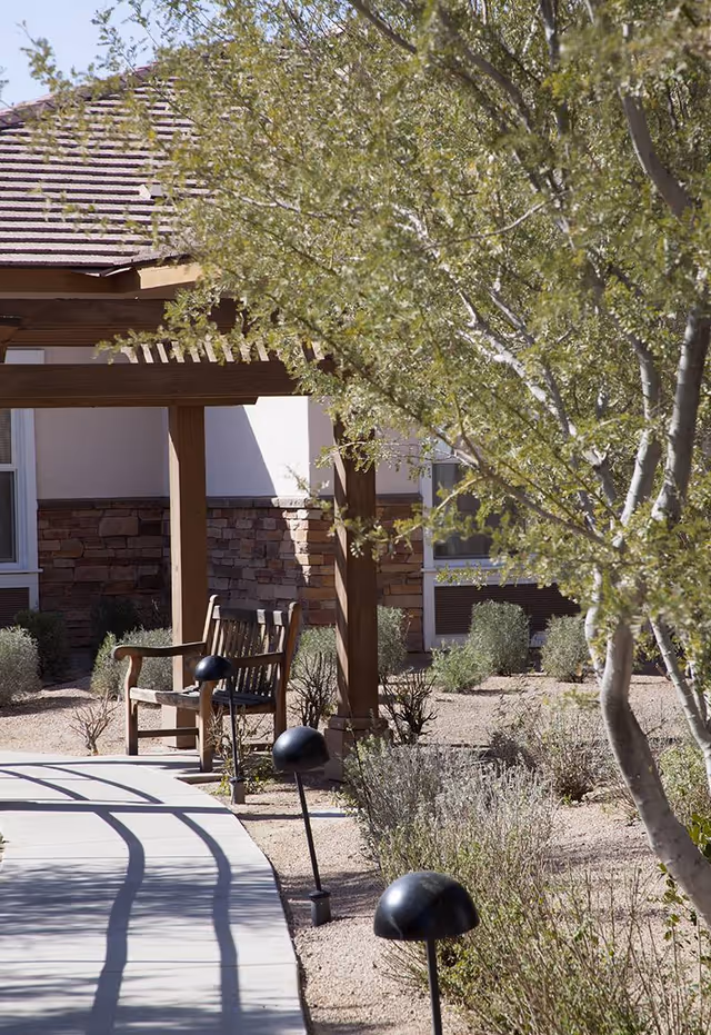 Landscaped outdoor walkway with low garden lights, a wooden bench under a pergola, and trees in front of a building.