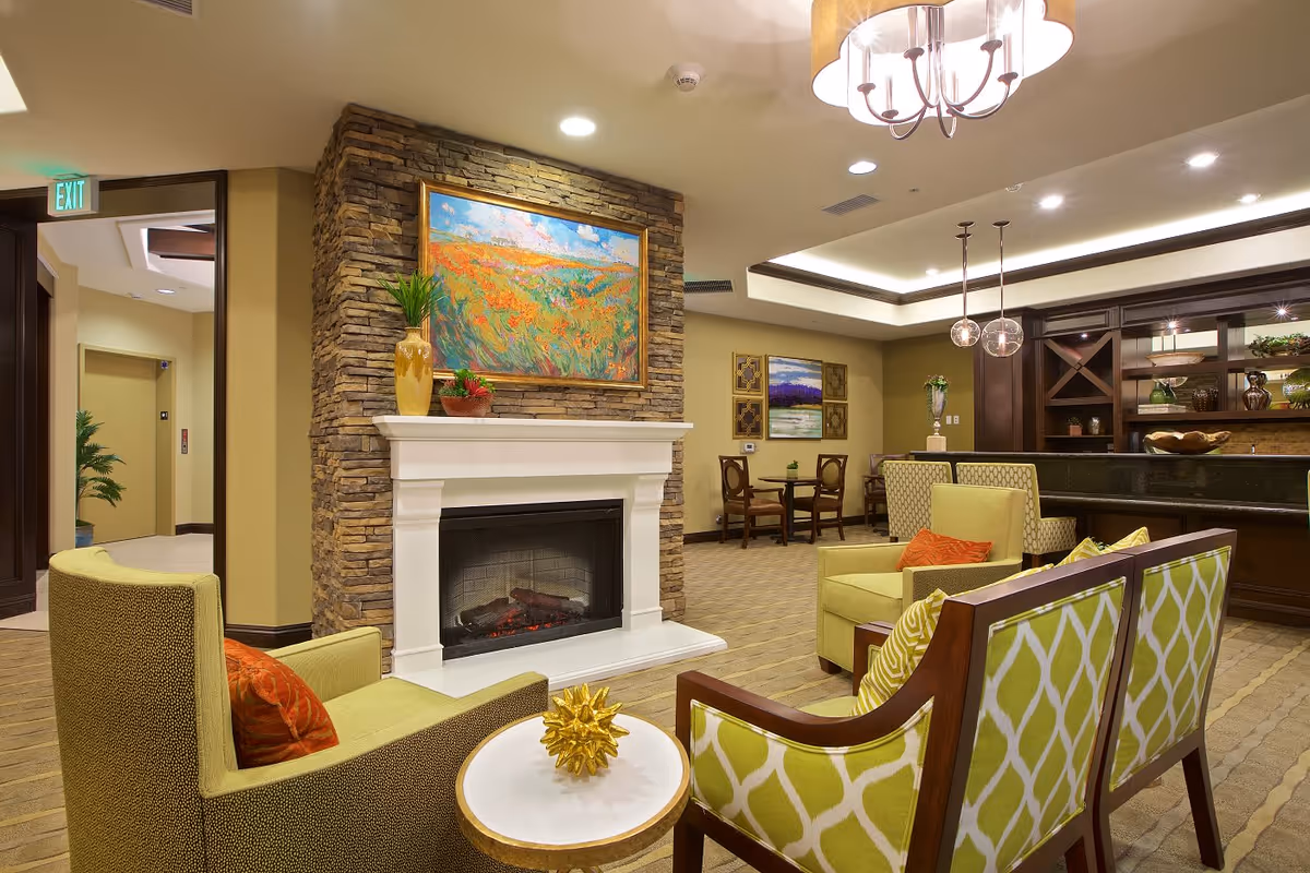 A cozy living room area in a senior living facility featuring a stone fireplace with a colorful landscape painting above it. The room has comfortable green and patterned chairs with orange and yellow cushions, a small round table with a decorative golden object, and a bar area with pendant lights and wooden cabinetry. There are additional seating areas and artwork on the walls, with warm lighting creating an inviting atmosphere.