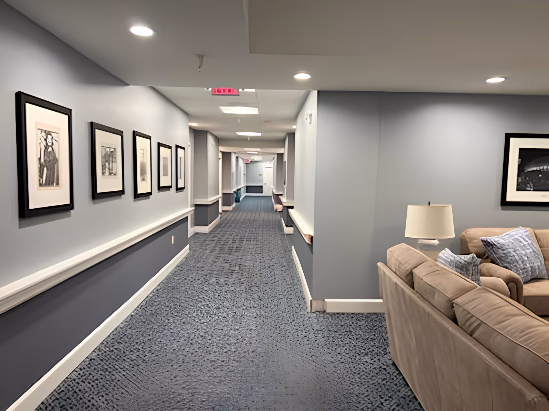 A long indoor hallway with blue carpet and gray walls adorned with framed black and white artwork. On the right side, there is a beige couch with blue and gray pillows and a table lamp. The hallway is well-lit with ceiling lights and has handrails along the walls.