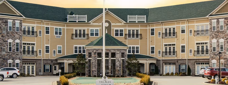 Front exterior view of The Villages at the River Club senior living facility, featuring a large three-story building with beige siding and stone accents, multiple windows and balconies, a central entrance with a covered portico, and a flagpole in front.