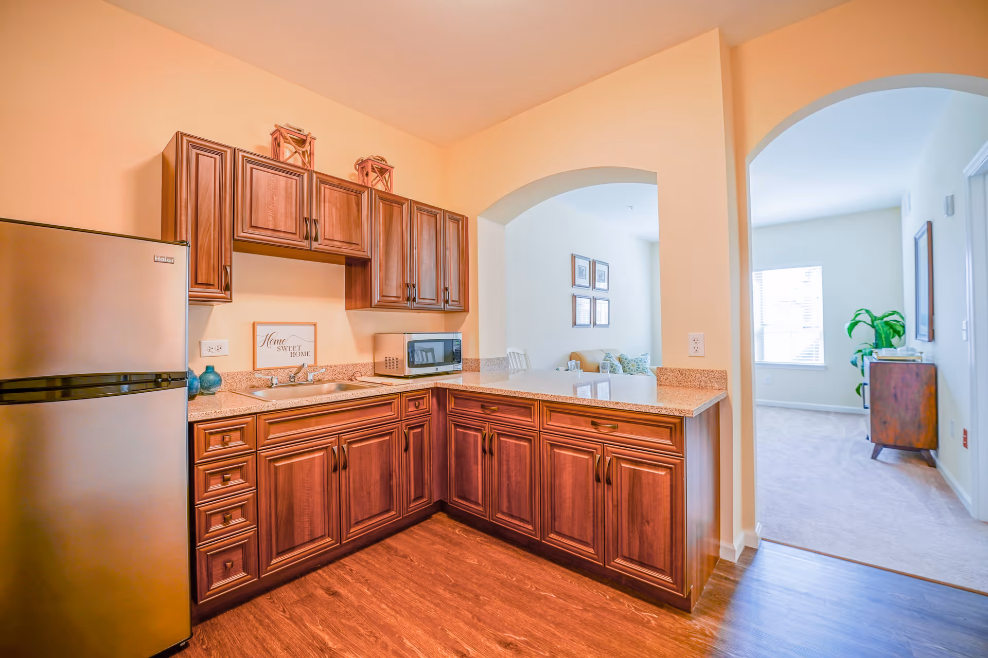 A kitchen area with wooden cabinets and drawers, a stainless steel refrigerator, a microwave on the countertop, and a small sign that reads 'Home Sweet Home'. The kitchen opens into a living room area with a window, a plant, and a wooden cabinet.