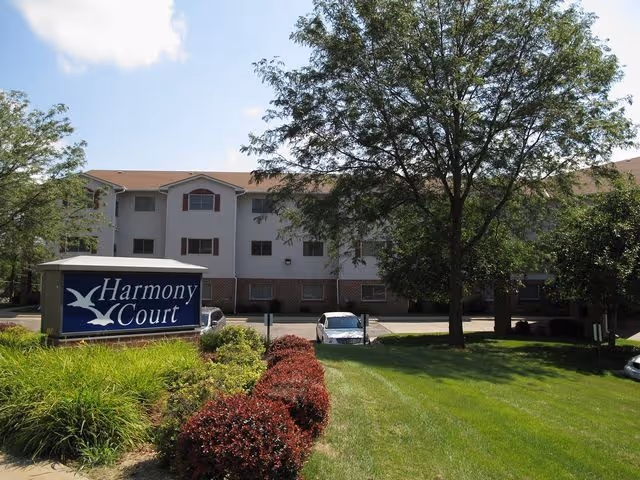 Front of a three-story Harmony Court senior living building with a lawn, trees, a sign, and parked cars.