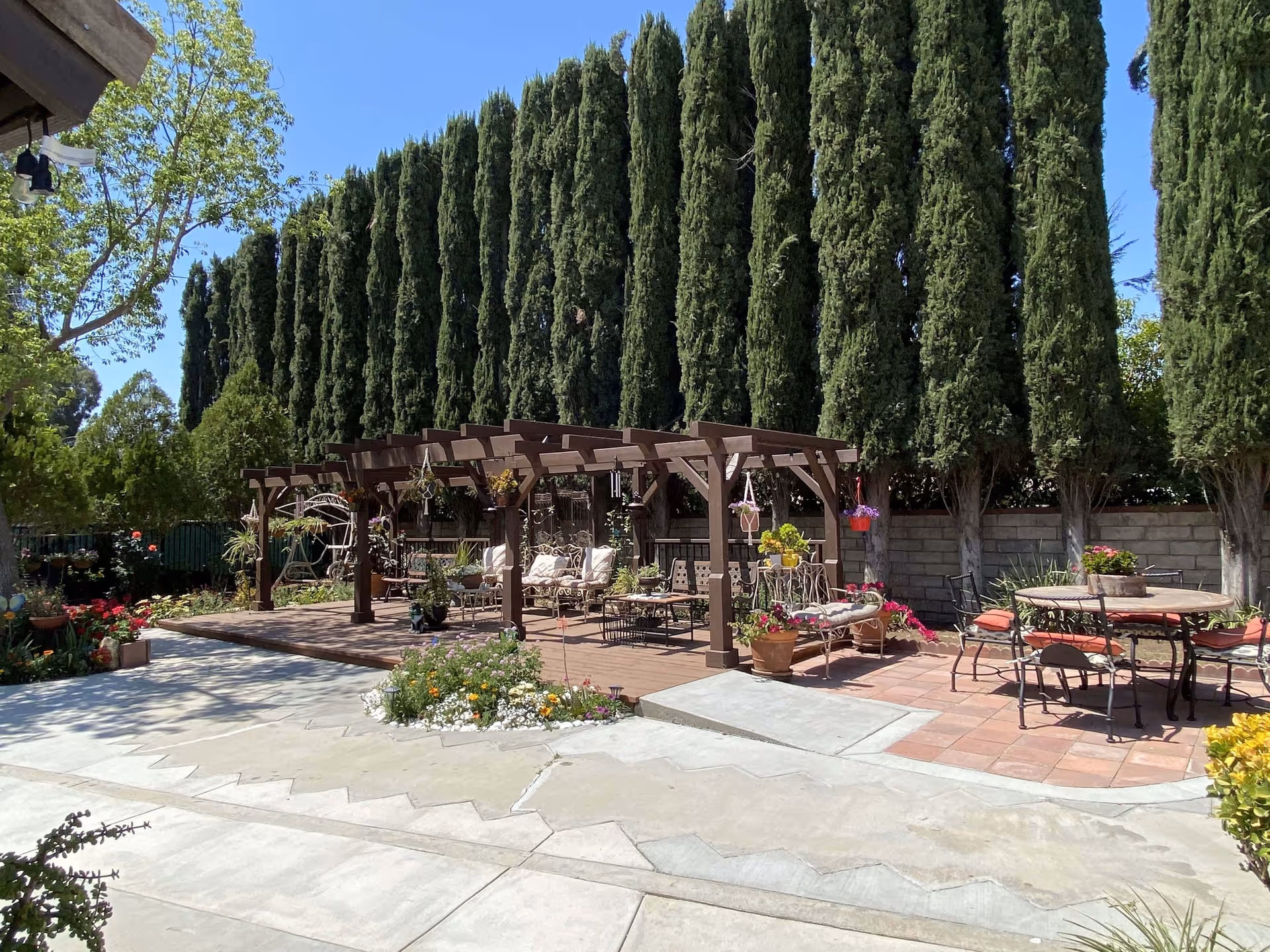 Outdoor patio area with a wooden pergola featuring cushioned seating and hanging plants, surrounded by tall evergreen trees and various flowering plants. There is also a round table with chairs on a tiled section of the patio under a clear blue sky.