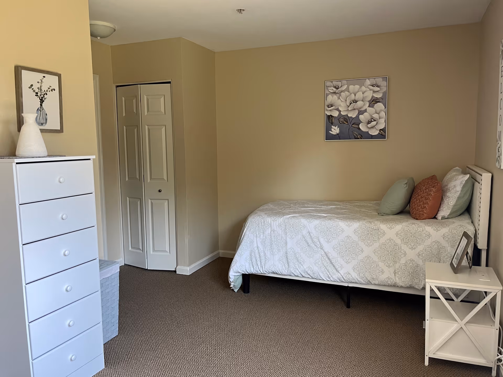 A simple bedroom with a single bed against the right wall, decorated with patterned bedding and three pillows in green, orange, and white. Above the bed is a framed floral artwork. To the left of the bed is a small white nightstand with a picture frame on it. On the left side of the room is a tall white dresser with five drawers, topped with a white vase and a framed picture of flowers. The walls are painted beige, and there is a closed white closet door in the corner. The floor is carpeted in a neutral tone.
