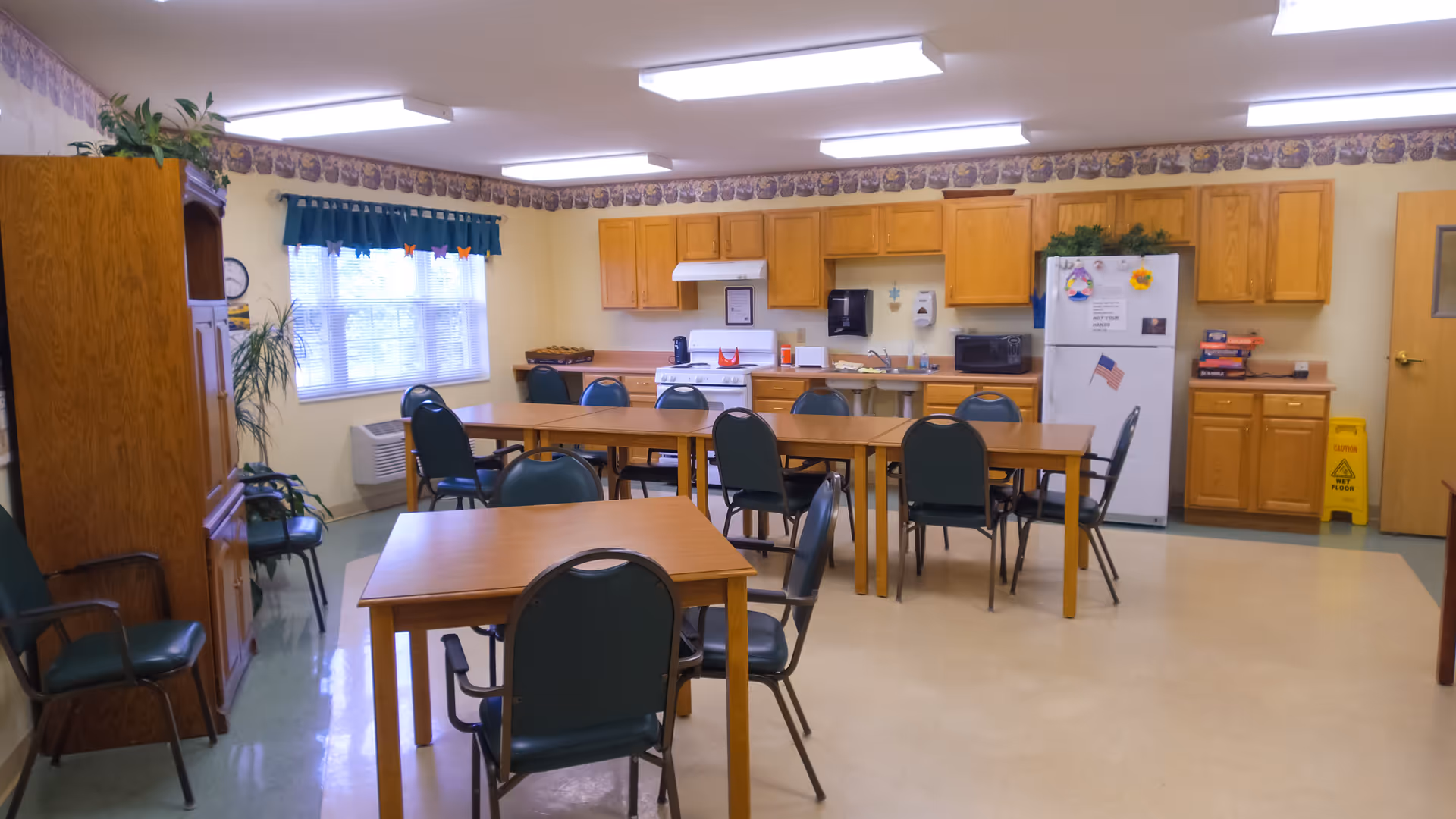 Community dining room with several wooden tables and green chairs facing a kitchenette with oak cabinets and a white refrigerator.