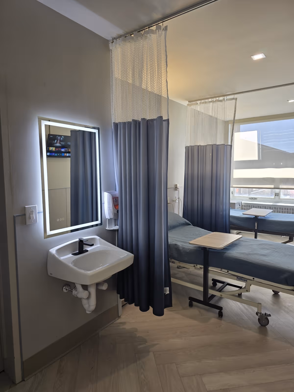 A partitioned patient room with two hospital beds, privacy curtains, a wall-mounted sink and illuminated mirror near a large window.