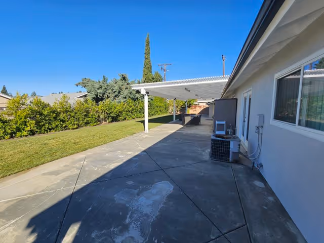 Outdoor patio area with a concrete floor and a white pergola attached to the side of a building. There is a grassy lawn with bushes and trees along the edge, and clear blue sky overhead.