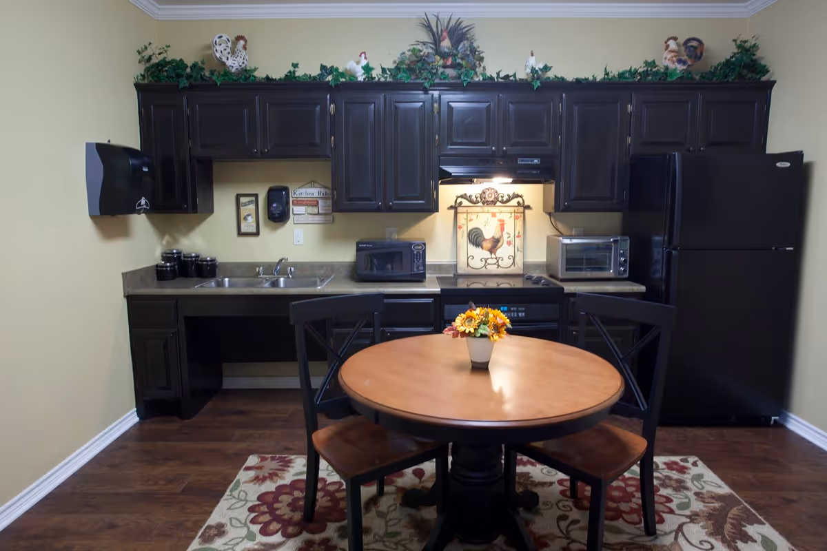 A small kitchen area with dark wooden cabinets, a double sink, a microwave, a toaster oven, and a black refrigerator. There is a round wooden table with two chairs in front of the kitchen, and a small flower arrangement on the table. The kitchen has decorative greenery and rooster figurines on top of the cabinets, and a floral rug on the wooden floor.