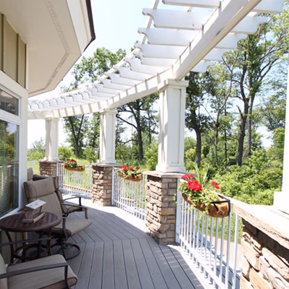 A bright outdoor patio area with cushioned chairs and a small round table. The patio features white pergola beams overhead and stone pillars with flower boxes containing red flowers attached to the white railing. Green trees and foliage are visible in the background.