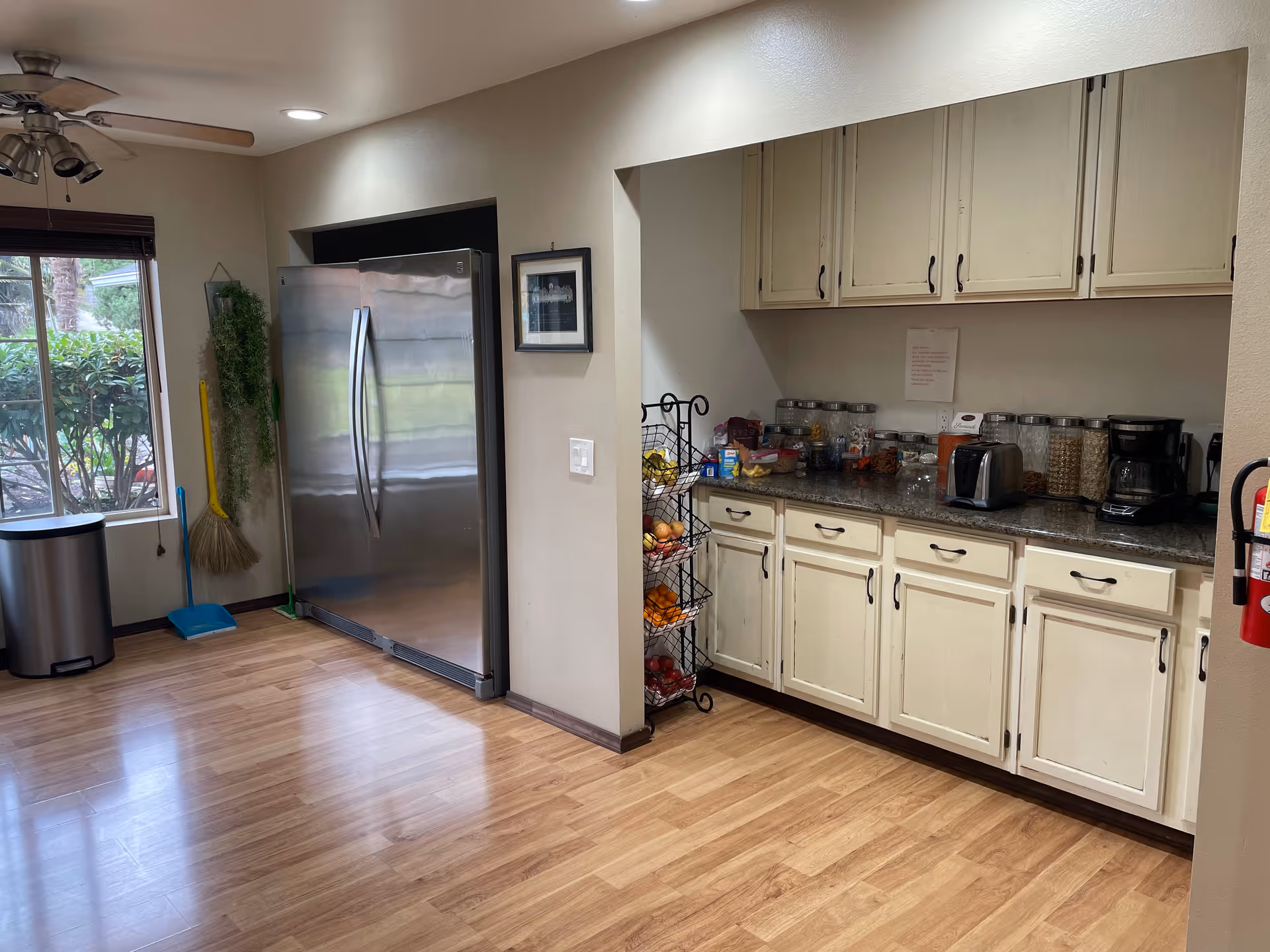 A kitchen area with wooden flooring, white cabinets, and a granite countertop. On the countertop, there are multiple jars filled with cereals and snacks, a toaster, and a coffee maker. To the left, there is a large stainless steel refrigerator next to a window with a view of greenery outside. Cleaning tools like a broom and dustpan are hanging on the wall near the refrigerator. A three-tiered fruit basket stand is placed beside the cabinets. A ceiling fan with lights is visible on the ceiling.