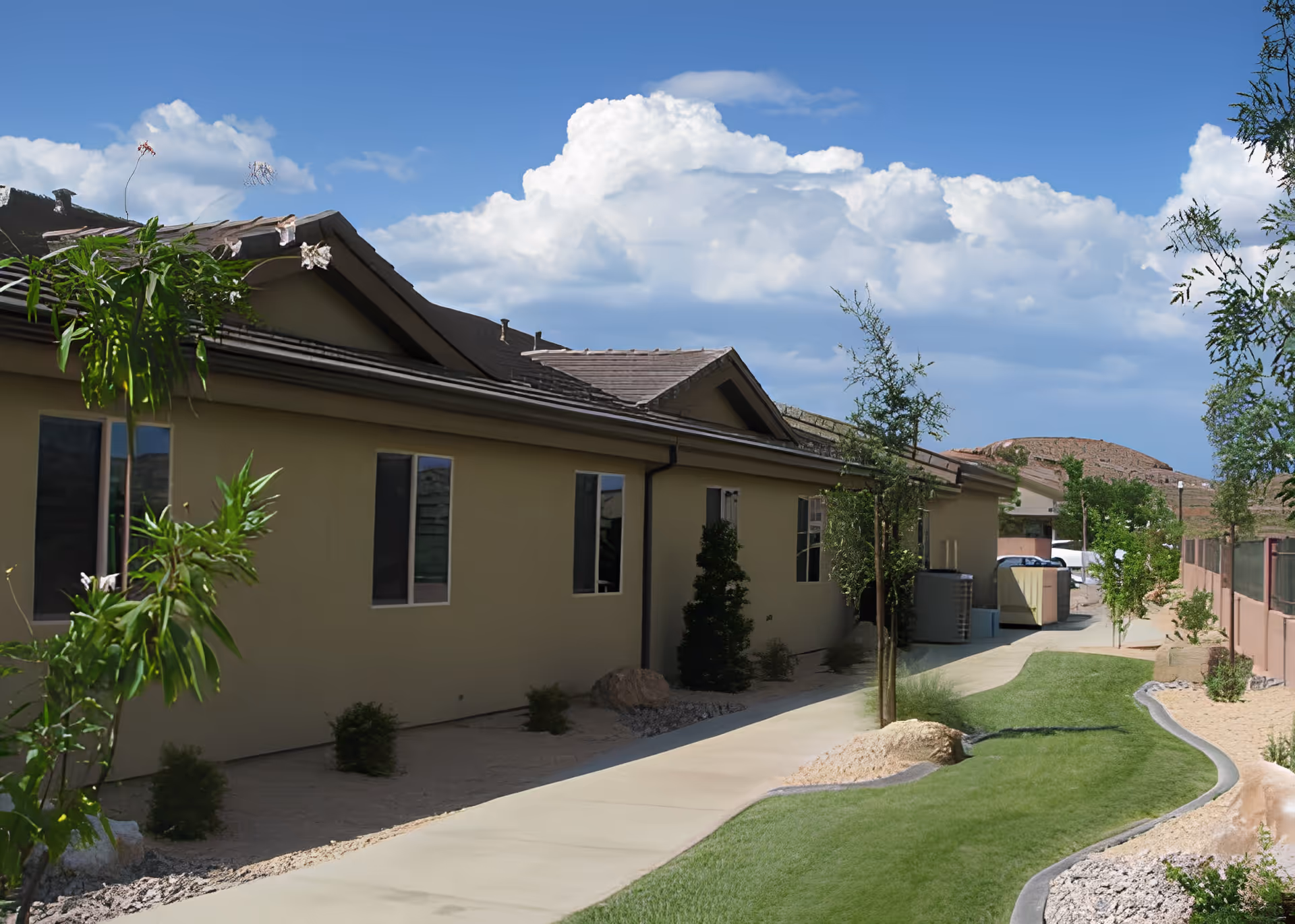 Side view of a single-story building with beige walls and a tiled roof, bordered by a paved walkway and landscaped with small trees, shrubs, and grass under a partly cloudy blue sky.