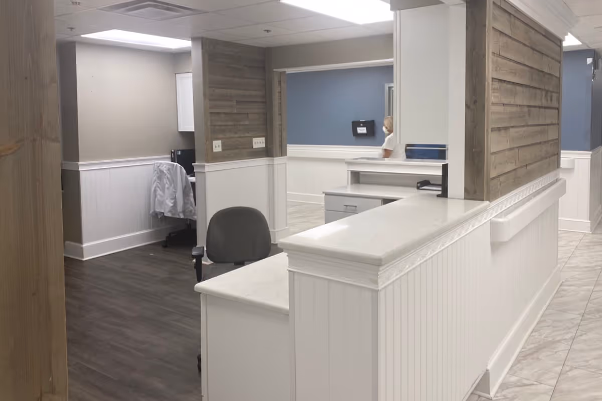 Interior view of a reception or nurse station area in a healthcare facility with white and wood-paneled walls, a white countertop, a gray office chair, and a person walking in the background wearing a mask.