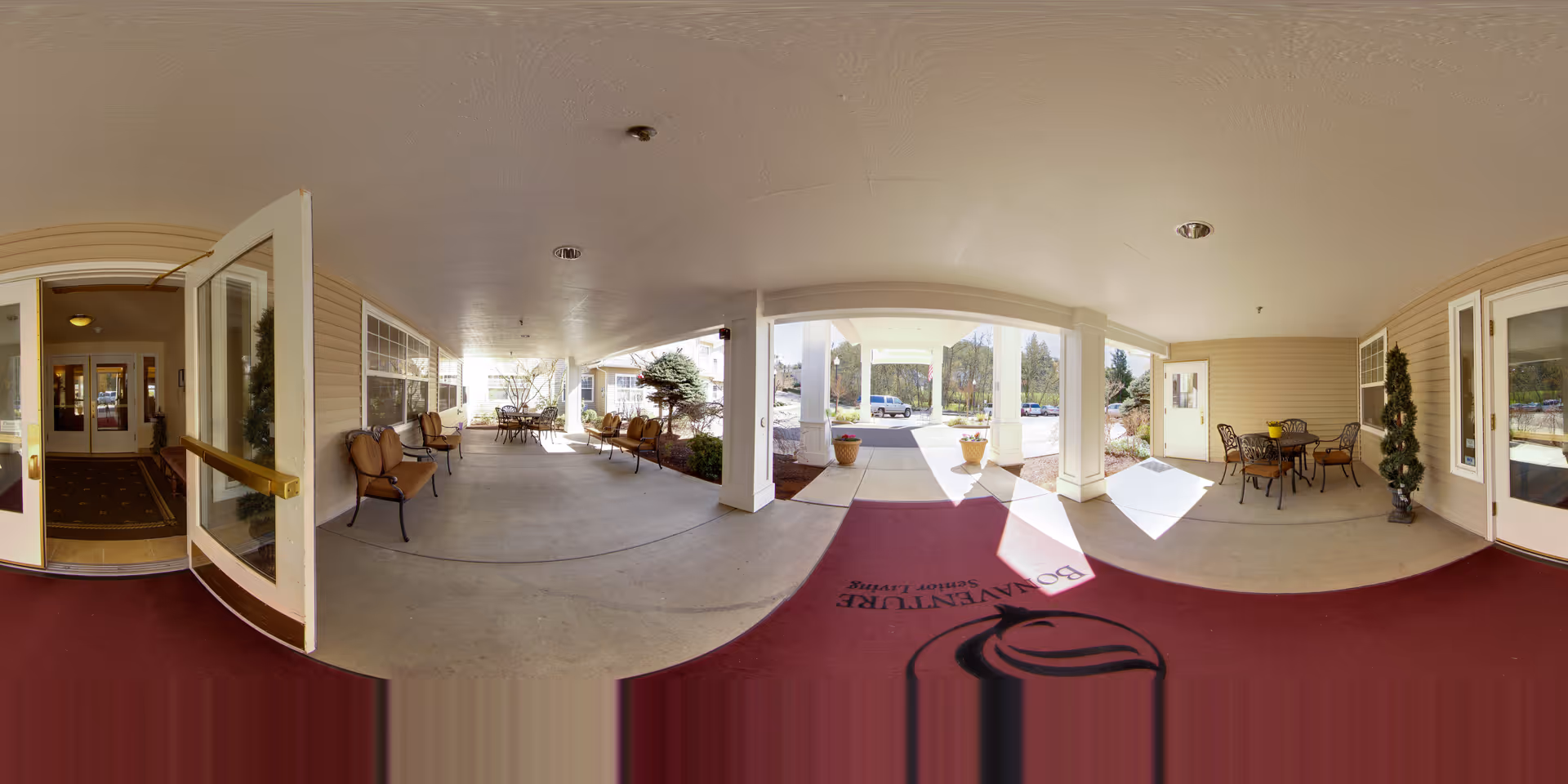 Covered outdoor entrance area of a senior living facility with seating arrangements including chairs and tables. The area has a maroon mat with the text 'Bonaventure Senior Living' and decorative plants near the walls. Doors lead inside the building, and there is a view of the parking lot and greenery outside.