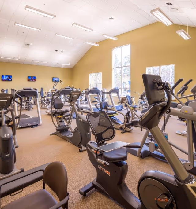 A bright fitness center with numerous treadmills, stationary and recumbent bikes and other exercise equipment under high ceilings and large windows.