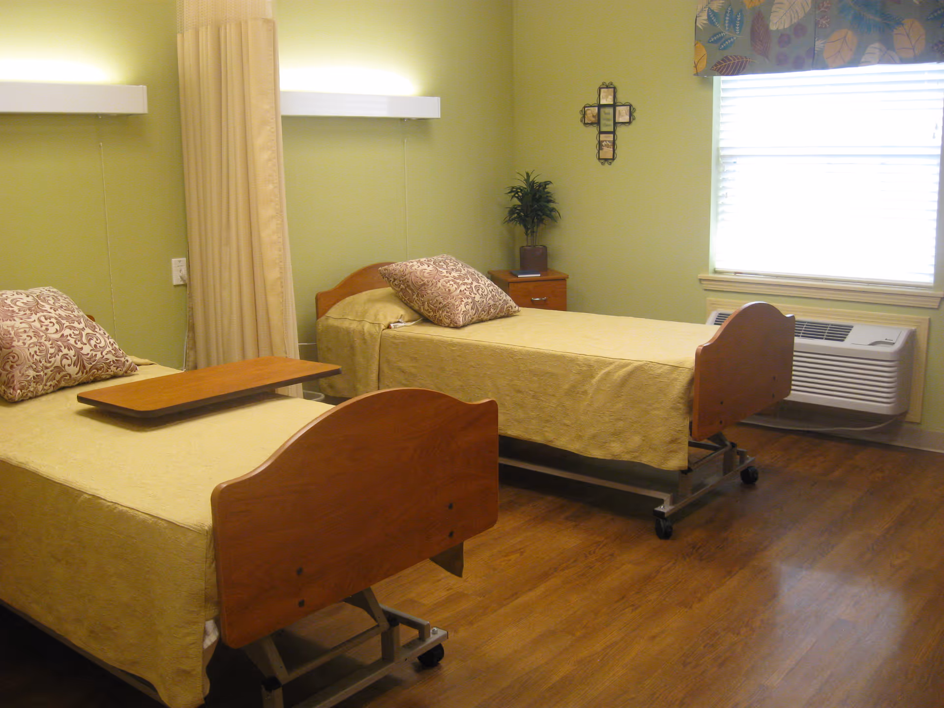 A nursing facility room with two single beds, each with a wooden headboard and footboard, yellow bedspreads, and patterned pillows. A small wooden bedside table with a potted plant and a decorative cross on the wall are visible. The room has light green walls, a window with blinds and a floral valance, and wood-look flooring.