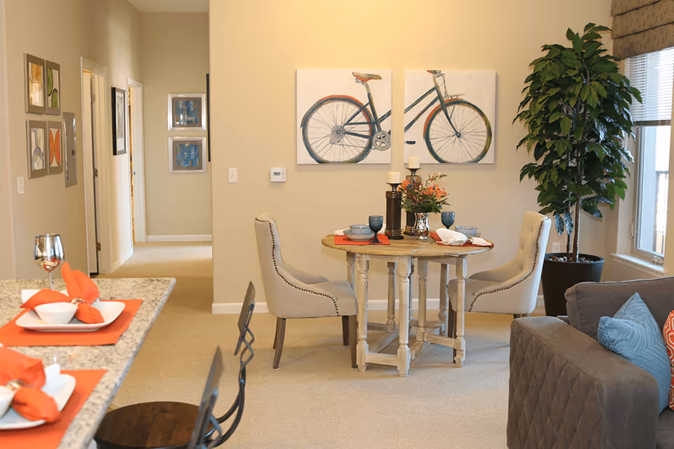 A cozy dining area in a senior living facility with a round wooden table set for four, featuring blue dishes, candles, and a flower arrangement. Two upholstered chairs are placed around the table. To the left, a kitchen counter with place settings and orange napkins is visible. A large potted plant stands near a window with blinds, and a gray couch with colorful pillows is partially visible on the right. The walls are decorated with artwork, including a two-panel painting of a bicycle.